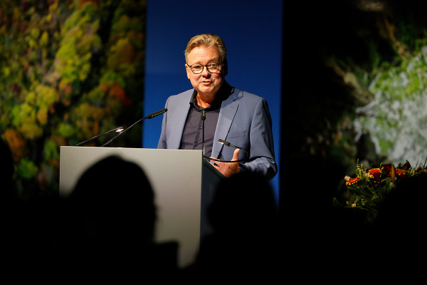 Rednerpult mit Thomas Berg im Zentrum. Im Hintergrund sind zwei Fotogarfien aus der aktuellen Gasometer-Ausstellung "Mythos Wald" zu erkennen.  unscharf zu erkennen