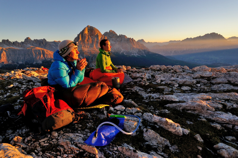 Ein Foto von zwei in dicken Jacken und Schlafsäcken auf Gestein sitzenden Personen beim Sonnenaufgang in den Dolomiten.
