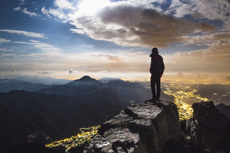 Foto von einem Wanderer auf einem Felsen mit Blick auf andere Berge und Lichter einer Stadt bei Dämmerung