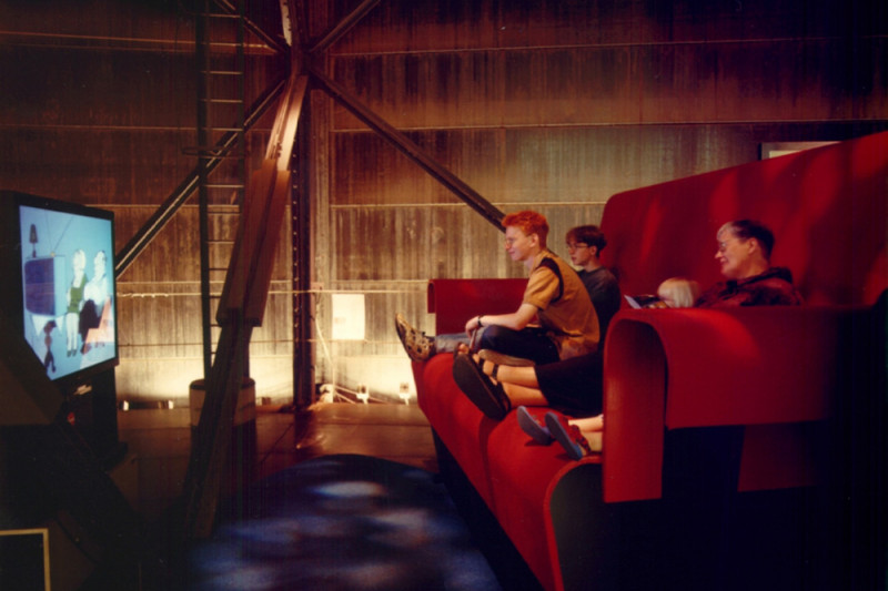 Drei Personen auf einem übergroßem rotem Sofa sitzend und auf den vor ihnen stehenden Fernseher schauenden.