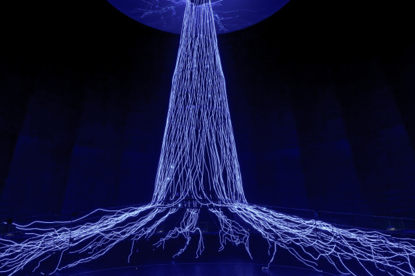 Leuchtende, 35 Meter hohe Lichtinstallation „Der Baum“ im Gasometer Oberhausen mit grün illuminiertem Stamm, ausladender Baumkrone und sichtbarem Wurzelwerk in einer dunklen Ausstellungshalle.