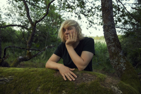 Dominik Eulberg lehnt an einem bemoosten Baumstamm in einem lichten Wald und blickt nachdenklich in die Kamera. Die natürliche Waldkulisse vermittelt Ruhe und Verbundenheit mit der Natur.